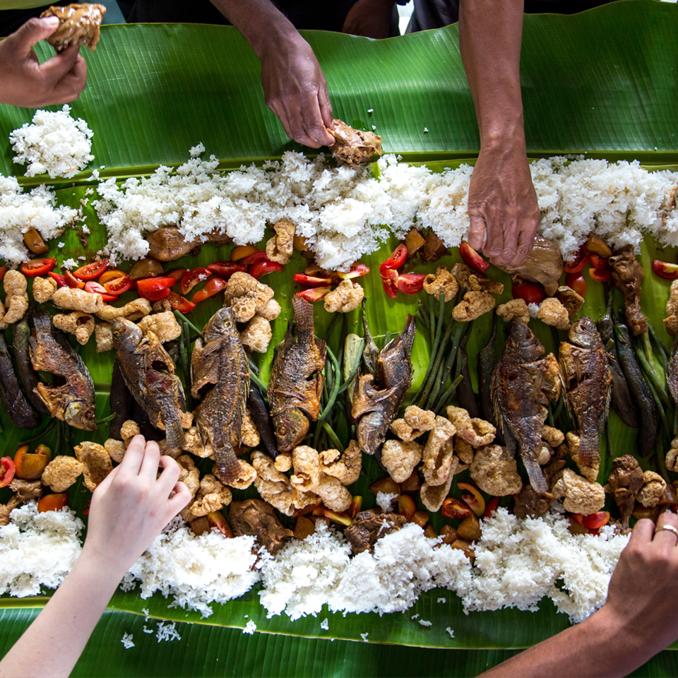 PEOPLE REACHING IN TO A MEAL OF FISH RICE AND TOMATOES