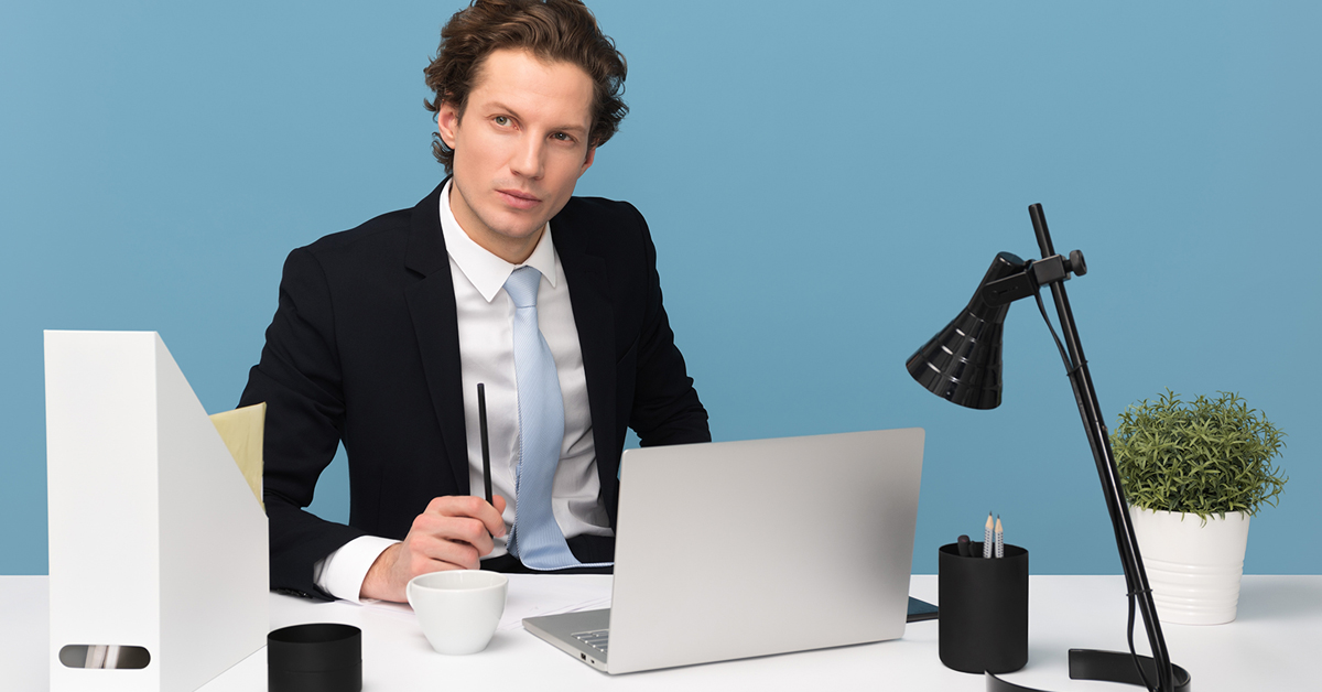 white male wearing a suit and light blue tie sitting at a desk with a computer